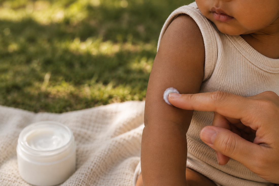 Close-up of a caregiver applying a small amount of moisturizer to the upper arm of a child with medium-brown skin while sitting outdoors on a soft cream blanket, with warm natural light and a blurred grassy background.