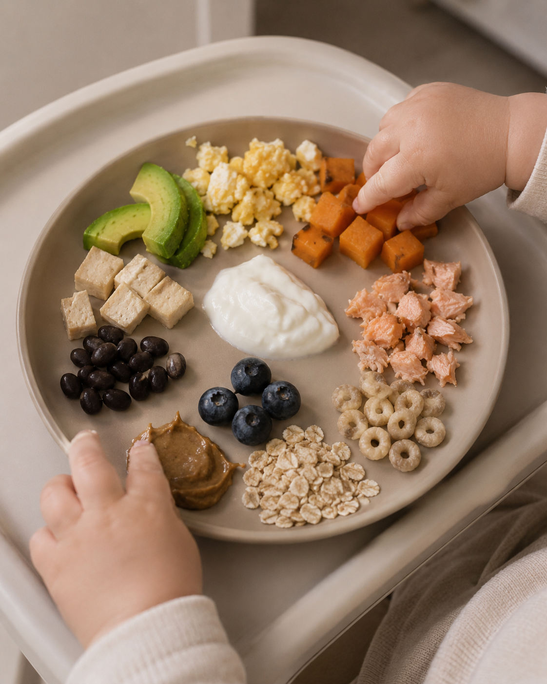 Baby hands reaching for a tan plate of baby-led weaning foods and common allergens including egg, yogurt, peanut butter, salmon, oats, beans, tofu, blueberries, avocado, and sweet potato on a high chair tray