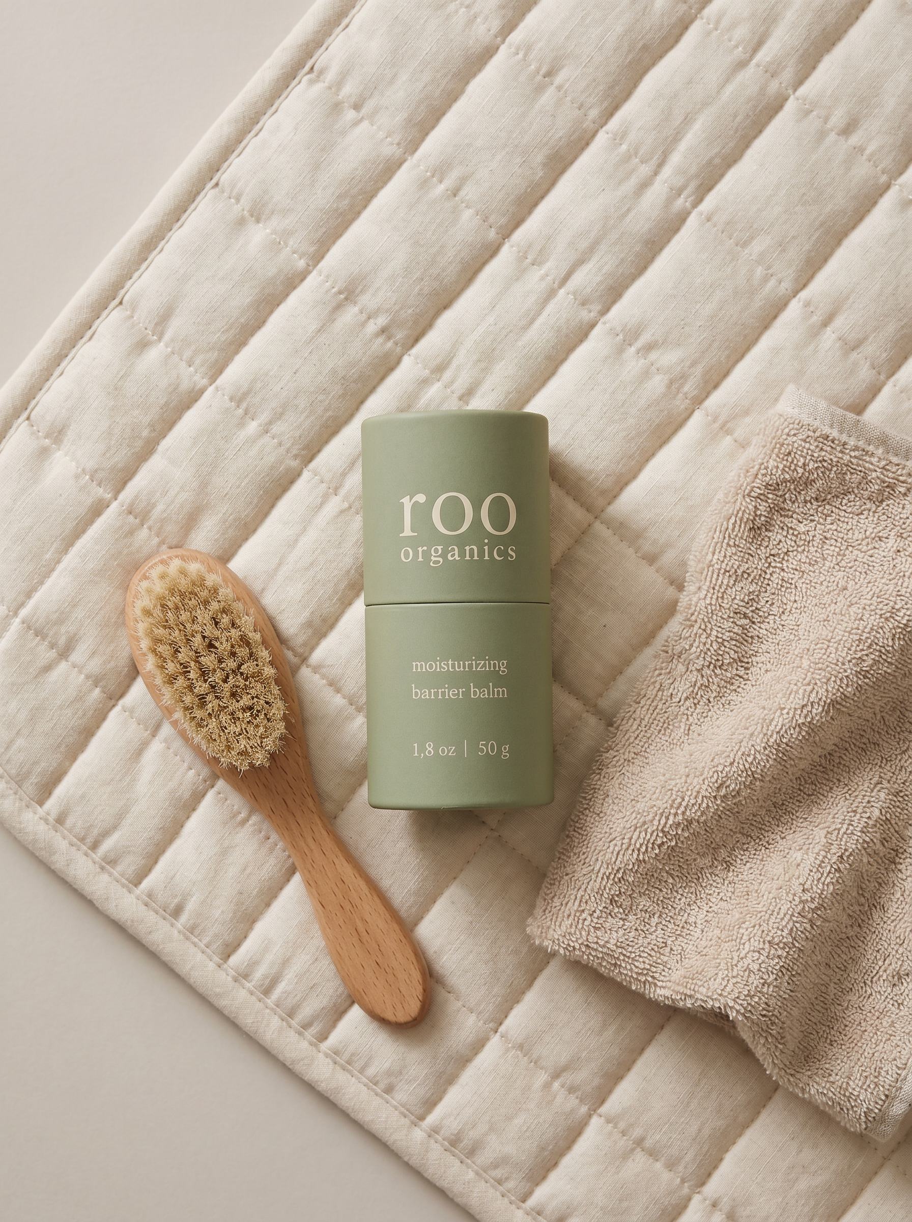 A green cylindrical container of Roo Organics moisturizing barrier balm placed on a soft quilted cream-colored surface, accompanied by a wooden baby brush with soft bristles and a folded beige towel.