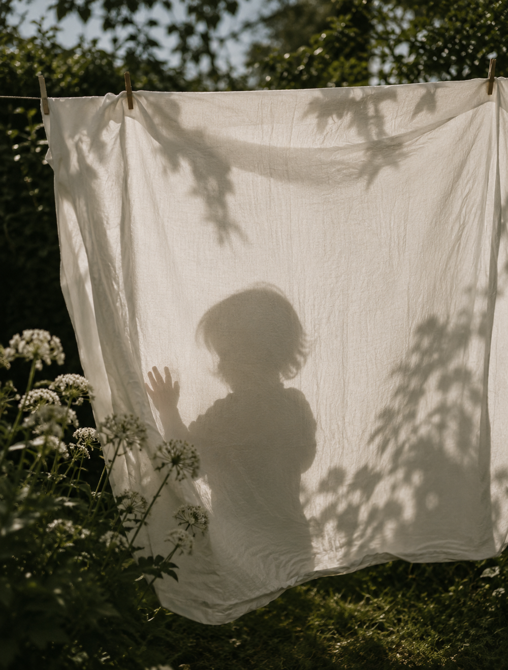 Silhouette of a child standing behind a light fabric sheet hanging outdoors, with shadows of leaves and plants visible on the sheet.